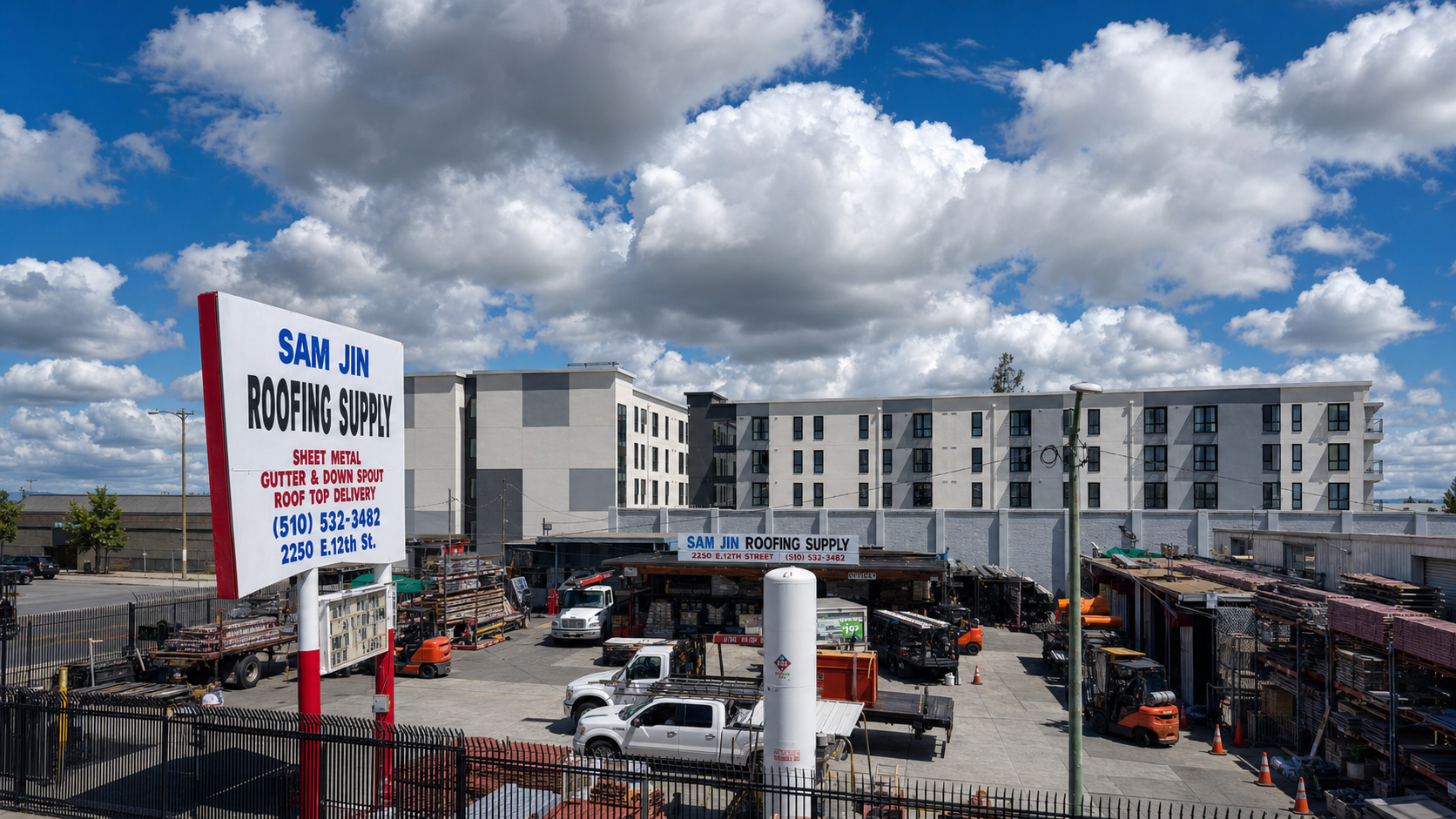 Sam Jin Roofing Supply yard at 2250 E. 12th Street in Oakland with the storefront sign, supply yard, trucks, and forklifts under a bright cloudy sky